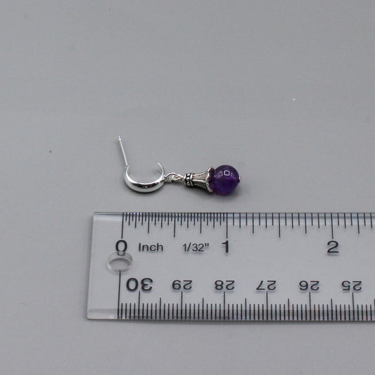 These expertly crafted earrings showcase the gentle sway of a vibrant amethyst gemstone, elegantly capped with a finely detailed pewter design. Suspended from a delicate silver hoop stud.