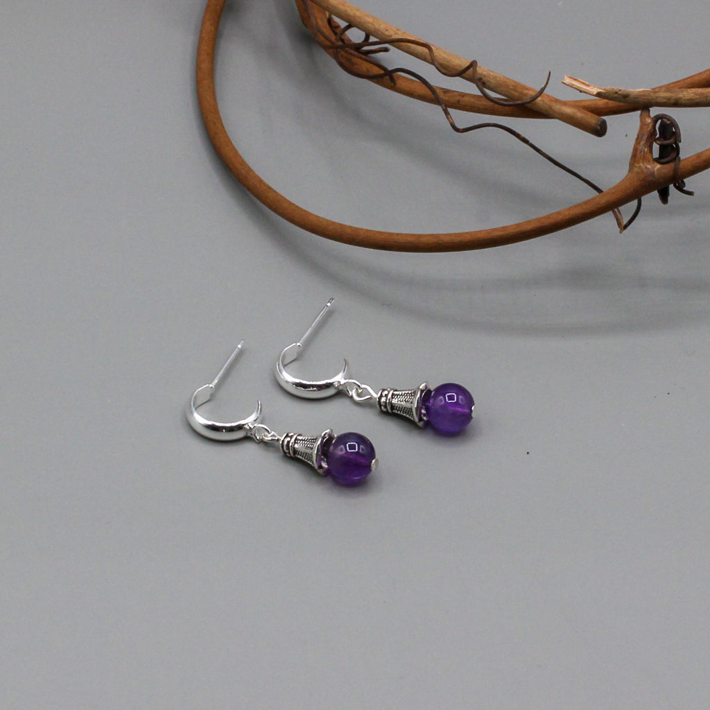 These expertly crafted earrings showcase the gentle sway of a vibrant amethyst gemstone, elegantly capped with a finely detailed pewter design. Suspended from a delicate silver hoop stud.