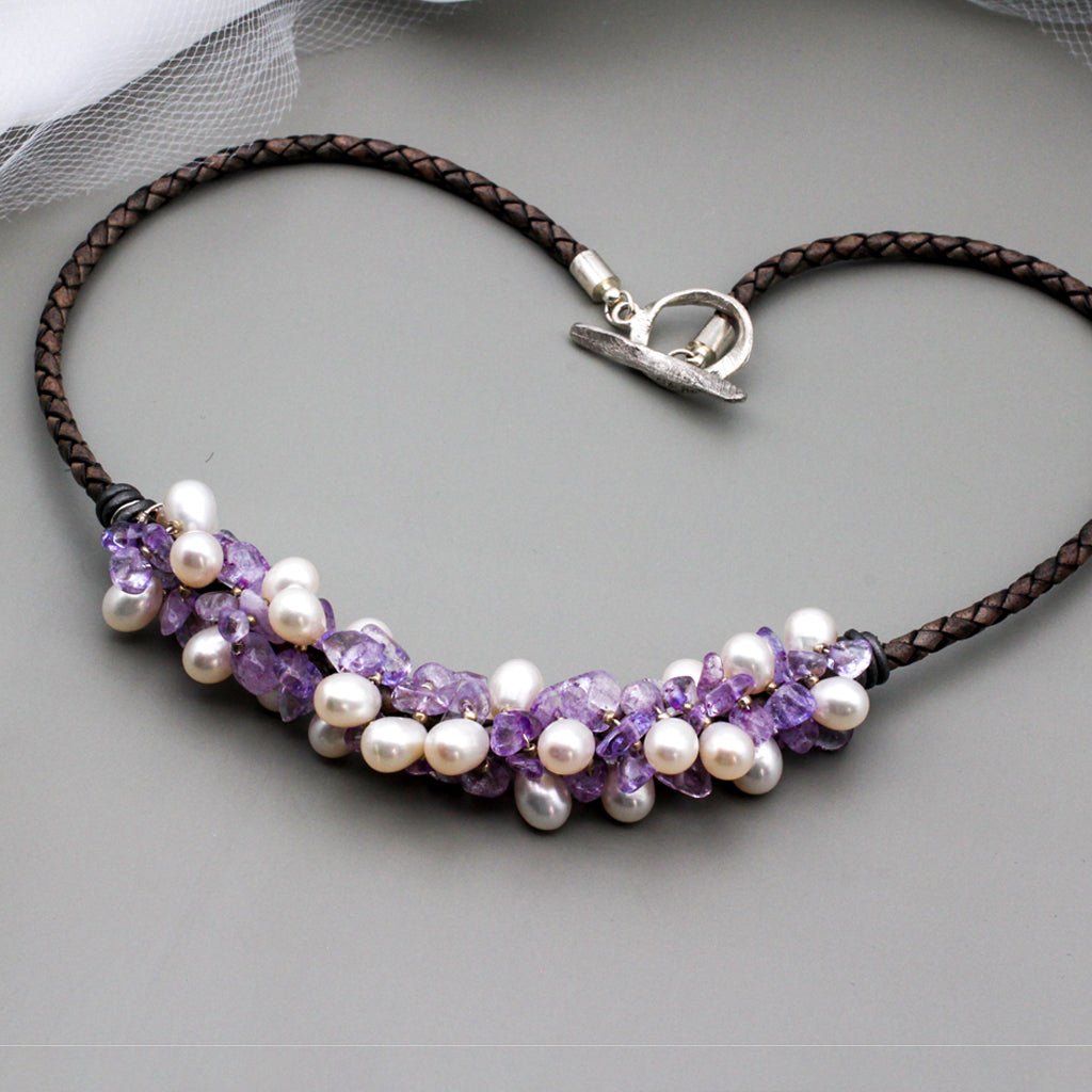 Amethyst gemstone beads and freshwater pearls twist around braided dark gray leather. It is finished with a silver toggle clasp.